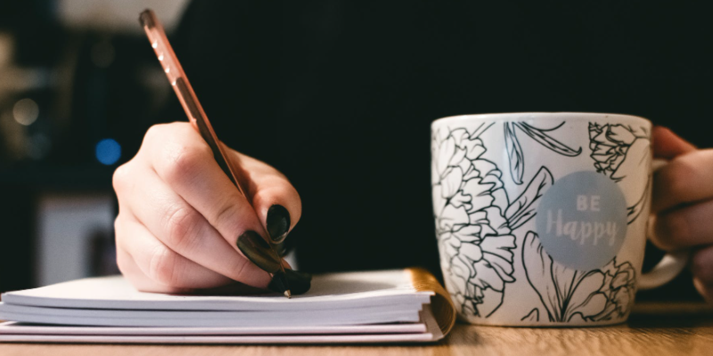imagem ampliada da mão de uma mulher escrevendo em caderno ao lado de uma xícara